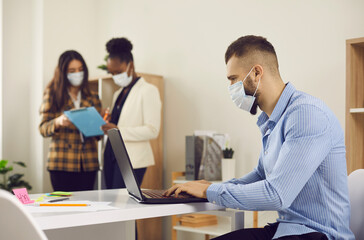 Man employee in face medical mask working on laptop indoors in office after lockdown with interracial colleague on background. New normal business practice of coronavirus covid-19 outbreak control