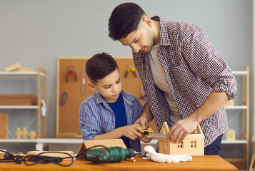 Adult man teaching little boy to work with wood. Serious child and father measuring wooden house with tape. Concentrated dad and son making toys at carpenter's workshop as part of DIY homework project
