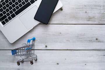 Top view of laptop, smartphone and cart on white wood background with copyspace for online shopping concept.