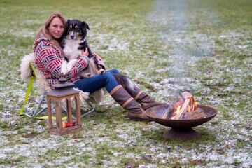 Blonde woman with her Australian Shepherd dog on her lap. In casual clothing, winterboots and thick winter sweater Outside on the snowy grass by the campfire in winter
