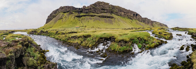 A gorgeous panoramic view of the landscape in Iceland. mountain river and a large mountain with a waterfall. Horizontal photography. Green meadows with grass. Postcard concept