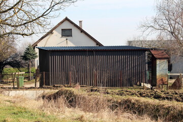 Elongated garage covered with dark corrugated metal protection in suburban family house backyard surrounded with rusted metal fence and uncut dry grass on cold sunny winter day