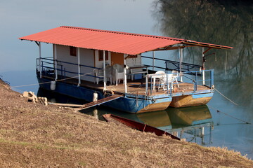 Fototapeta na wymiar River barge converted into river boat house with improvised homemade roof and terrace with white plastic chairs and tables left at local river bank covered with dry grass on cold sunny winter day