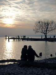 Loving couple watches the sunset on Lake Garda. Figures in silhouette stroll on a small pier. Lovers smoke a cigarette sitting on a log.