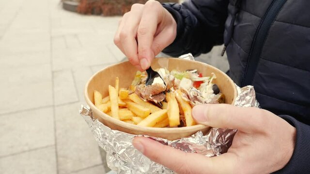 Man on a street sit eating takeaway food box with kebab meat with salad and fried potatoe chips from disposable tableware