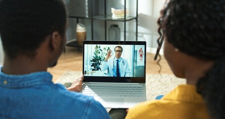 Rear of African American couple man and woman sitting in living room in house and videochatting on...