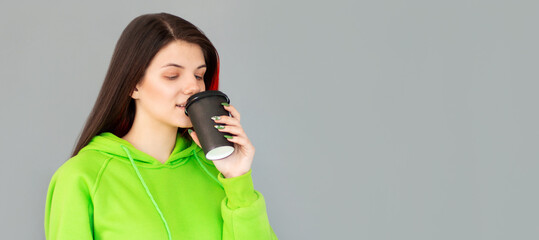 Studio shot of girl with holds paper cup of hot coffee, wears green jumper, grey background. Beautiful young woman enjoys aromatic cappuccino.