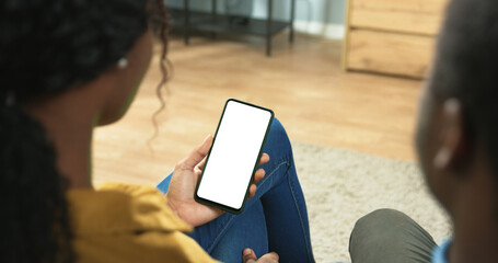 Close up of African American couple man and woman sitting in room surfing on smartphone with green...