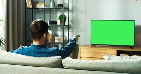 Rear of Caucasian young guy watching TV with chroma key resting on sofa sitting in living room in...