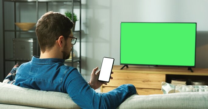 Caucasian man in glasses sitting on sofa in modern room in house watching TV with green screen and typing on smartphone with chroma key browsing online. Back view, leisure concept
