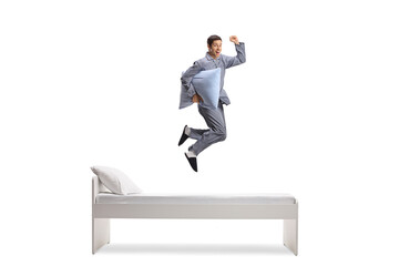 Full length shot of a man in pajamas holding a pillow and jumping on a bed