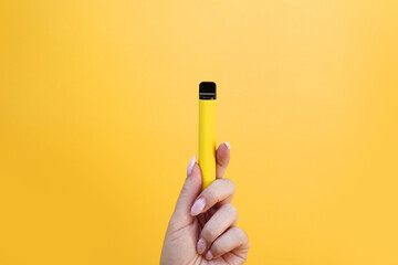 Yellow disposable electronic cigarette in female hand. Bright yellow background