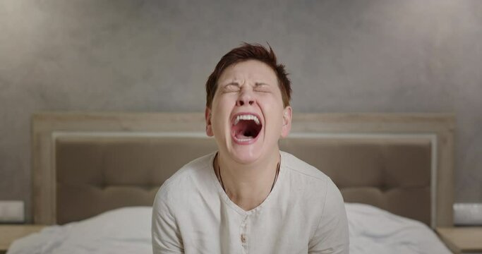 Mature woman screaming in despair. Unhappy middle aged female with short hair clenching fists and shouting with closed eyes while sitting on bed at home