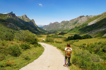 A girl with a backpack is looking to the mountains and there is a blue sky and some white clouds