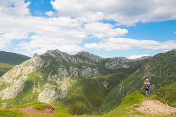 A girl with a backpack is looking to the mountains and there is a blue sky and some white clouds