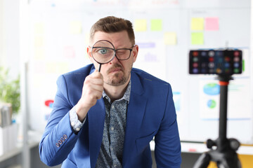 Young business coach holding magnifying glass near eye in front of mobile phone camera