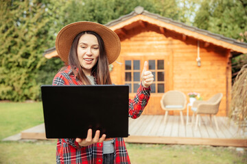 Young gardener shows her wooden garden shed on a laptop. Woman with a straw hat is standing in the...