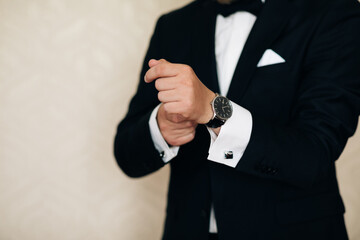 Businessman adjusts his white shirt button. Concept successful businessman. Portrait of trendy attractive stunning man in black tuxedo with tie fasten button on sleeve cuffs of his white shirt.