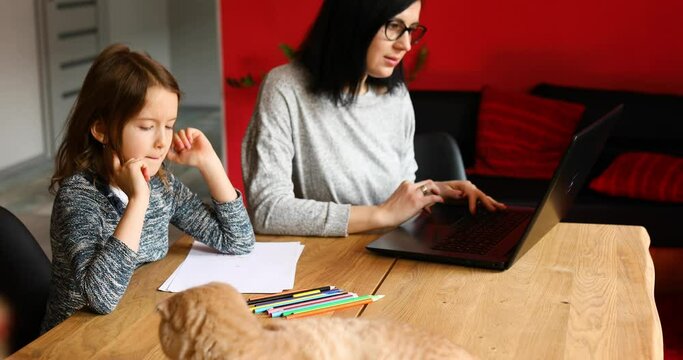 Mother working in her home office on a laptop, her daughter sits next to her and draw, Scottish cat sitting on the table too. Woman freelance, remote work and raising a child at workplace.