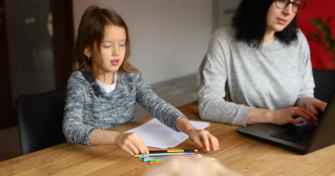Mother working in her home office on a laptop, her daughter sits next to her and draw, Scottish cat sitting on the table too. Woman freelance, remote work and raising a child at workplace.