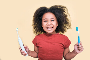 Smiling African American little girl holds manual and electric sonic toothbrushes, isolated on beige background