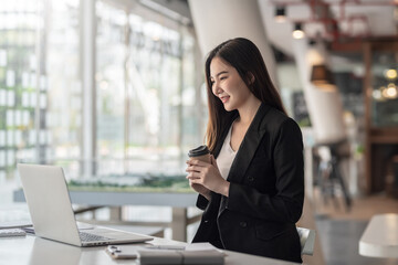 Side view of a young Asian businesswoman holding a coffee mug looking at the laptop at office.