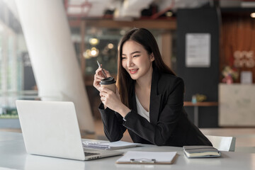 Young asian businesswoman holding a coffee mug is working on a laptop at office.
