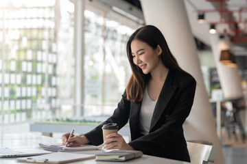 Beautiful smiling asian businesswoman holding a coffee cup while taking notes at office.