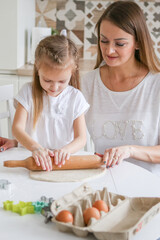 Happy mom and daughter prepare dough together using rolling pin, smiling young mom, charming girl prepare cookies, buns in the kitchen. The concept of a friendly family preparing for Easter