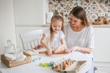 Happy mom and daughter prepare dough together using rolling pin, smiling young mom, charming girl prepare cookies, buns in the kitchen. The concept of a friendly family preparing for Easter