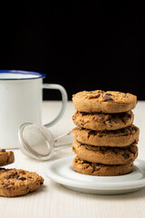Close-up of stack of chocolate cookies in white plate and white cup, on white wooden table, black background, in vertical, with copy space