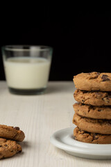 Close-up of stack of chocolate cookies and glass of milk, selective focus, on white wooden table, black background, vertical, with copy space