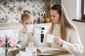 Loving young mother in a sweater is talking to a cute funny baby daughter enjoying time together at home in the kitchen with tea. happy family. mom with little baby girl fun play feel joy hugging 