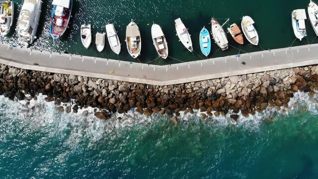 Small fishing boats and tourist yachts moored at shelter, small port of Hersonissos town, top-down aerial shot at sunny day. Sea waves break on rough stones from other side of seawall