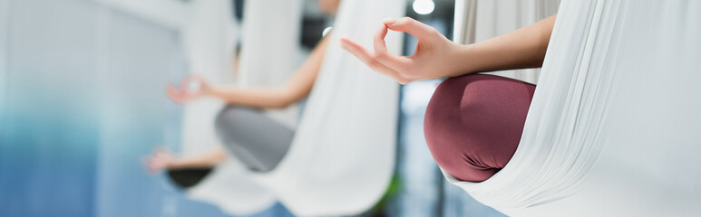 cropped view of women meditating in lotus pose in aerial yoga hammocks, blurred background, banner