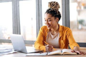 Online education, e-learning. Happy african american young woman in stylish casual clothes, studying remotely, using a laptop, listening to online lecture, taking notes while sitting at home, smiles - obrazy, fototapety, plakaty