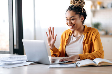 Online video communication. Joyful attractive African American woman, freelancer or student, in casual clothes, uses laptop for online video conference with colleagues or friends, greets, smiles