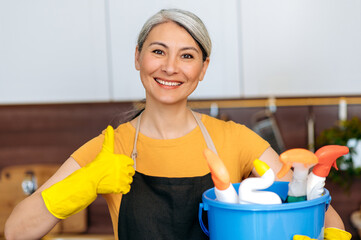 Cleaning service concept. Portrait of happy senior gray-haired Asian cleaning lady or housewife, wearing apron and gloves, holding bucket, shows thumb up, looks at camera, smiling, ready for cleaning
