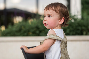 A baby 1-2 years old with a scratch on his forehead looks to the side, looking for his mother, against the background of a green bush. Childhood. 