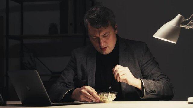 Night work. Late dinner. Corporate discipline. Overtime schedule. Sneaky starving male employee enjoying eating food at dark office workplace with laptop.