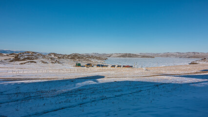 Small village on the shore of the lake. Several wooden houses with green roofs. Lake Baikal is covered with ice, hills on the banks. Shadows on the snowy land