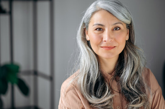 Close-up portrait of beautiful influential confident mature gray-haired Asian woman ceo, lawyer, real estate agent, stands in the office, dressed in stylish clothes, looks at camera, friendly smiling