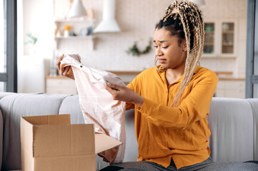 A confused sad african american girl unpacking her parcel at home. Surprised young woman got an...