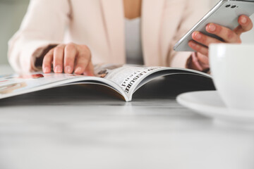 Woman with mobile phone reading magazine at white table, closeup