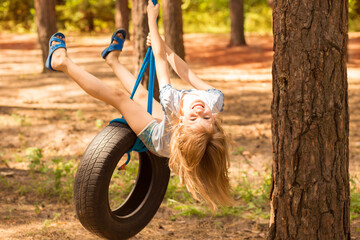 Cute little girl swinging on wheel attached to big tree in autumn forest. 