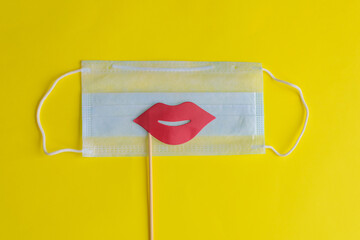 Medical face mask, paper pink lips on stick on bright yellow background, close-up. Celebrating April Fools ' Day during coronavirus pandemic concept.