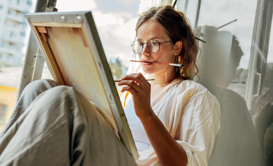 Closeup portrait of a pretty female artist painting on canvas in her art studio sitting next to the window. A woman painter with eyeglasses painting with oil searching for imagination in the workshop. - obrazy, fototapety, plakaty