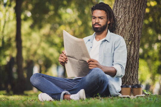 Full size photo of serious afro american man sit grass park relax read newspaper article in outdoors outside