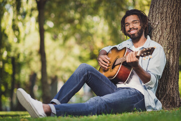 Portrait of attractive cheerful guy sitting on green grass playing guitar singing romantic hit...
