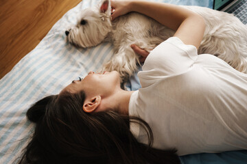 Happy white woman stroking her dog while lying on bed at home